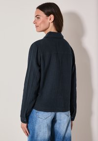 Černá texturovaná košile s límečkem, dlouhými rukávy a knoflíkovými manžetami, doplněná světle modrými džínovými kalhotami. Zpětný pohled ukazuje detaily švů.