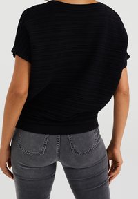 Černý texturovaný top s krátkým rukávem a žebrovaným vzorem, spárovaný se šedými džíny z rifloviny, které mají zadní kapsy a mírné vyblednutí.