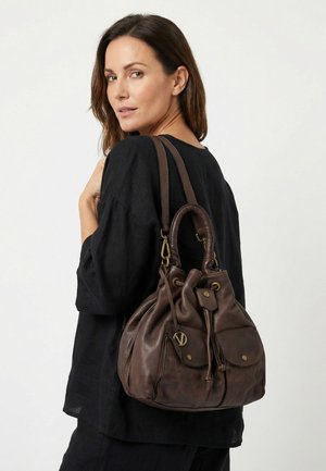 Žena oblečená v černém drží přes rameno velkou hnědou koženou kabelku se šňůrkovým zavíráním a předními kapsami, mírně otočená dozadu.