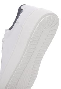 Suela de zapatilla blanca con un patrón de agarre texturizado, con un talón visible y costuras laterales, capturada desde un ángulo bajo sobre un fondo blanco.