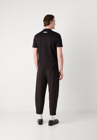 Fekete, rövid ujjú, slim fit póló, kicsi hátsó címkével. Laza fekete nadrággal és fekete cipővel, fehér zokni felett.