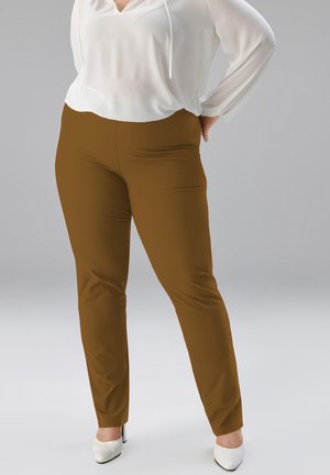 Frau trägt braune, eng anliegende Hose, weiße Bluse mit Kordelzug und weiße High Heels, steht mit einer Hand in der Hüfte.