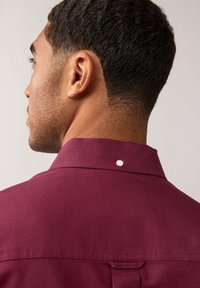 Camicia bordeaux con colletto a bottoni, realizzata in tessuto texturizzato e un singolo bottone bianco sul retro. Il design presenta dettagli di cucitura sottili.