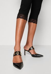 Sorte lædermules med spidse tæer og stiletto hæle. Har en bred rem med runde guldfarvede detaljer. Sorte leggings med blondekant.