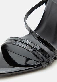 Κοντινή λήψη στο μπροστινό μέρος ενός μαύρου σανδαλιού από πατινέ δέρμα με δύο λεπτές παράλληλες λουρίδες πάνω από τα δάχτυλα και μια λοξή λουρίδα πάνω από αυτές.