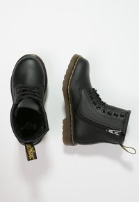 Paire de bottes en cuir noir à lacets avec fermetures éclair latérales, coutures jaunes et semelles en caoutchouc marron sur fond blanc.