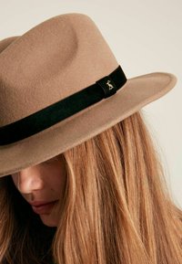 Beige felt hat with a black velvet band featuring a gold accent. Wide brim and soft texture, angled downwards. Close-up view.