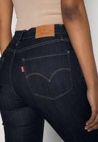 Osoba v tmavě modrých džínsech Levi's a béžovém topu, ukazující zadní kapsu s červenou nálepkou Levi's a hnědým koženým brandovým štítkem.