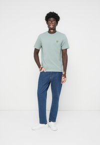 T-shirt en coton vert clair avec un petit logo jaune sur la poitrine, associé à un jean en denim bleu et des baskets blanches. Coupe décontractée, manches courtes.