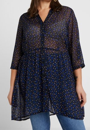 Puoliläpinäkyvä tummansininen tunika, jossa pieni valkoisen ja violetin geometrinen kuvio, etupuolella napit, kolmivartiset hihat, puettu sinisten farkkujen päälle.