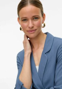 Šviesiai mėlynas švarkas su glotnia tekstūra ir išpjautomis atlapomis, derinamas su dideliais auksiniais žiedais. Modelis turi natūralų įvaizdį su tvarkingais plaukais.