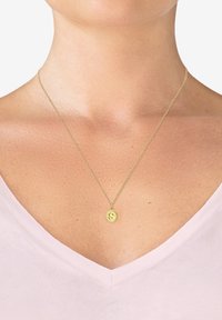 Gul vedhæng halskæde med cirkulært charm, der har en præget 'S', på en delikat kæde. Båret over en lys pink V-hals top.