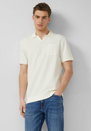 Jonge man die een witte korte mouwen polo met een borstzak en een blauwe spijkerbroek draagt, terwijl hij tegen een lichtgrijze muur staat.