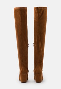 Botas marrones de ante por encima de la rodilla con una textura suave, parte superior ancha y tacones cuadrados. Los acentos laterales incluyen un pequeño detalle de lazo.