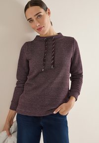 Kobieta w długim rękawie strukturalnej purpurowej bluzie z troczkami, w ciemnoniebieskich spodniach, trzymająca jasnoszary kurtkę, opierająca się o beżową ścianę.