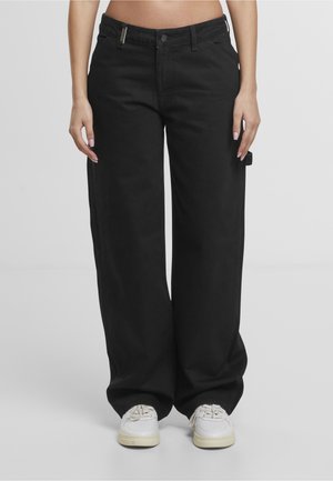 Personne portant un pantalon noir ample et des baskets blanches, debout sur un fond uni gris clair.
