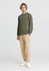 Felpa verde oliva con colletto rotondo e spalle cadenti, abbinata a pantaloni della tuta beige a sigaretta e sneakers bianche. Tessuto dalla texture liscia.