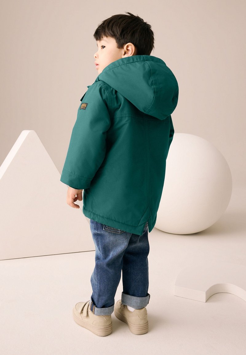 Dieťa v zelenej kapucňovej bunde, modrých džínsoch a béžových topánkach stojí a pozerá sa doľava na neutrálne pozadie s geometrickými tvarmi.
