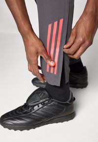 Pantalones deportivos grises con acentos de tres rayas rojas, ajustados por una mano, combinados con tacos negros y calcetas negras.