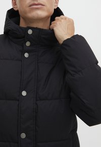 Doudoune noire avec un col haut, dotée de boutons-pression métalliques et d'une texture matelassée, mettant en avant la chaleur et l'isolation.
