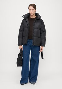 Čierna prešívaná bunda s vysokým golierom, zipsovým predným zapínaním a cvokmi. V kombinácii s hnedým sveterom a širokými džínsami.