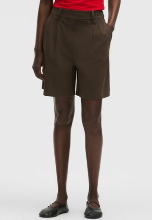 Donkerbruine op maat gemaakte knielange shorts gedragen met een rode top en zwarte platte schoenen door een staande persoon tegen een witte achtergrond.