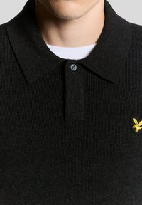 Czarna prążkowana koszulka polo z kołnierzykiem, z jednym guzikiem przy szyi i małym żółtym logo orła na piersi.