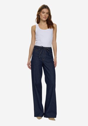 Femeie purtând un top alb fără mâneci și blugi evazați largi, cu talie înaltă, de culoare albastru închis, legați în talie, stând în picioare pe un fundal simplu.