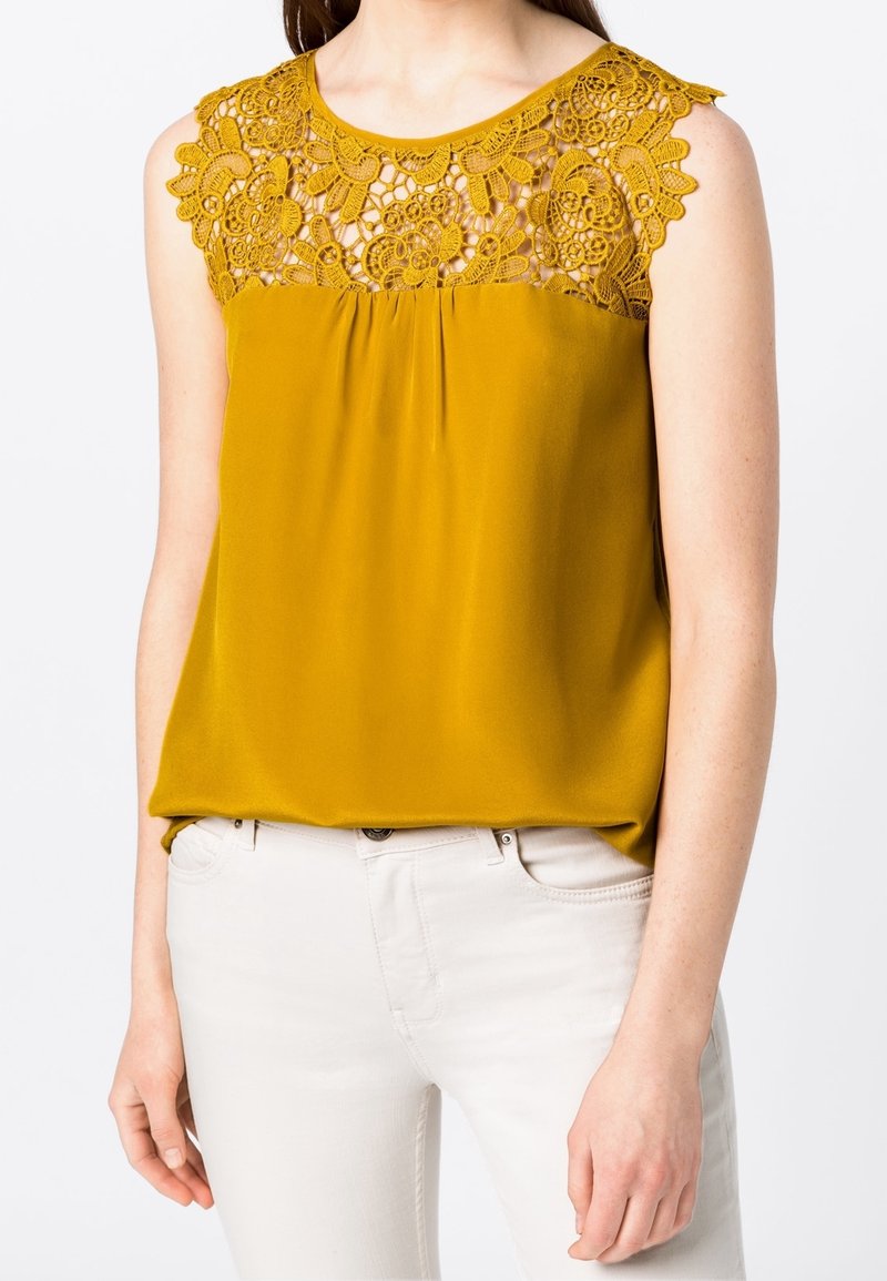 Blouse jaune moutarde avec un empiècement en dentelle orné de motifs floraux. Tissu doux et lisse avec une coupe décontractée, associée à un pantalon de couleur claire.