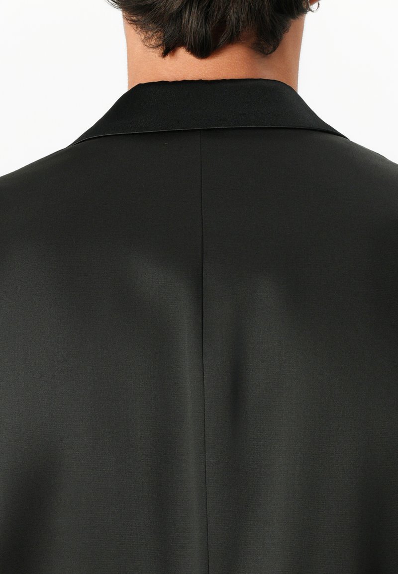 Veste noire courte avec col et couture au dos, portée par une personne avec les cheveux courts et foncés, vue de dos sur fond blanc.