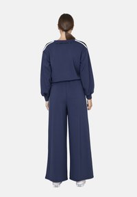 Survêtement bleu marine avec un pantalon à jambes larges et un haut ample. Présente des bandes latérales blanches et le logo "Marc Ellis" dans le dos.