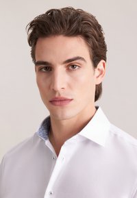 Biała koszula z kołnierzykiem w niebieski wzór paisley, gładka w dotyku, zapinana na guziki. Charakteryzuje się dopasowanym krojem i subtelnym połyskiem.