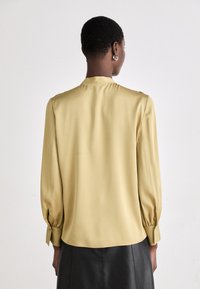 Blusa a maniche lunghe in morbido tessuto giallo chiaro con una texture liscia, caratterizzata da un colletto sottile e polsini arricciati. Abbinata a una gonna nera.