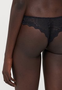 String en dentelle noire avec motif floral, bords festonnés et texture transparente. Conçu pour une couverture minimale avec une apparence délicate.