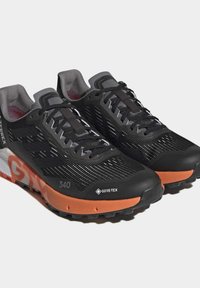 Mustaa juoksukengät, joissa on teksturoitu päällinen, valkoisella raidalla ja "GORE-TEX" -logolla. Oranssit yksityiskohdat pohjassa. Kevyt muotoilu.