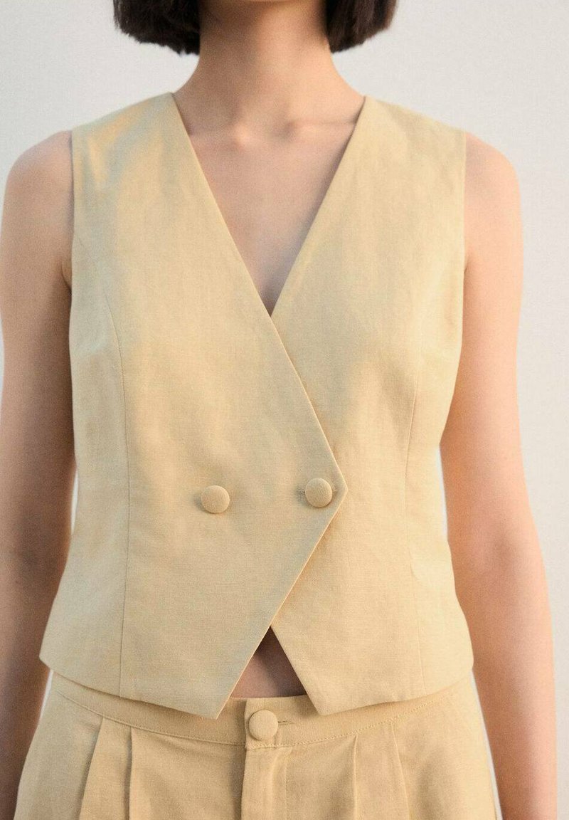 Gilet smanicato avvolgente beige con scollo a V, caratterizzato da due bottoni abbinati e tessuto strutturato. Abbinato a pantaloni beige plissettati.