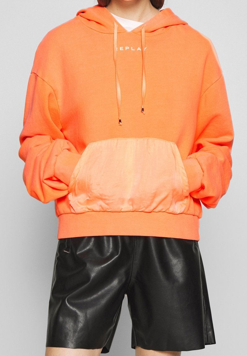 Sweat à capuche orange avec cordons de serrage et poche avant en tissu soyeux. Présente une coupe décontractée, des manches longues et un ourlet côtelé. Porté par-dessus un short en cuir noir.