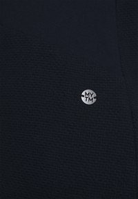 Marinblått textil med en slät sektion och en rund silvertagg med "MYTM"-logotyp fäst.