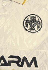 Vit tröja med ett geometriskt mönster. Har en svart cirkulär logotyp och stora svarta texter. Gula detaljer på kragen ger extra tydlighet.