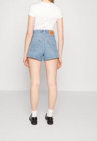 Light blue denim shorts with a high waist, frayed hem, back pockets, and a leather patch at the waistband. Styled with black shoes and white socks.