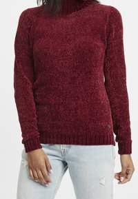 Pull à col roulé bordeaux avec une texture côtelée, une coupe ajustée et des poignets côtelés, arborant un petit logo au niveau de l'ourlet.