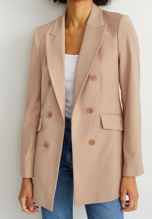 Person iført en beige dobbelttradet blazer over en hvid top og blå jeans, stående mod en ensfarvet lys baggrund.