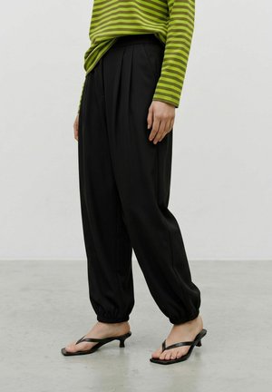 Personne portant un pantalon noir à ourlet froncé, un haut à manches longues rayé vert et noir, et des sandales noires à talon kitten sur un sol gris.