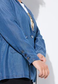 Blå denimjacka med långa ärmar, featuring silverknappar på ärmsluten och sidofickor, som visar en lätt tygtextur.