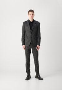Koolgrijs op maat gemaakt pak in een geweven stof, met een blazer met één knoop en bijpassende broek, gecombineerd met een zwart overhemd en schoenen.