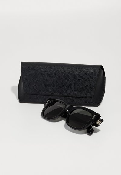 Schwarze Sonnenbrille mit quadratischem Rahmen, begleitet von einem strukturierten schwarzen Etui mit geprägtem Branding.