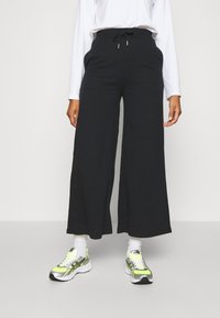 Svarta vida byxor med dragsko i midjan och sidofickor, tillsammans med sneakers med neondetaljer och vita ankelsockor.