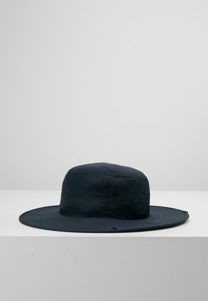 Chapeau de soleil noir à large bord avec couronne ronde, détails cousus et œillets de ventilation, posé sur une surface blanche devant un fond gris uni.