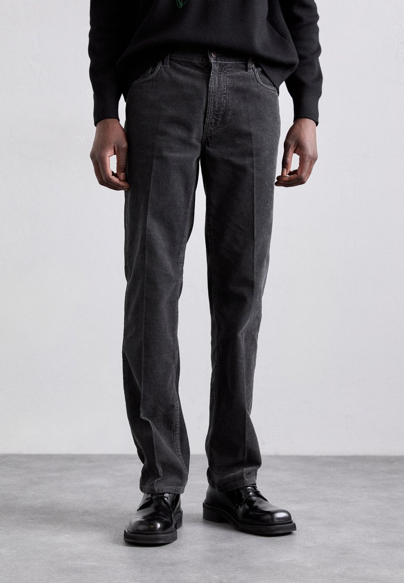 Black, straight-leg jeans with a textured finish, featuring a classic waistband, five pockets, and subtle stitching details. Worn with black shoes.
