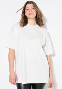 Bílý oversized tričko vyrobené z bavlny, s krátkými rukávy a jemným oranžovým nápisem "YOU" na přední straně. Hladká textura, volný střih.